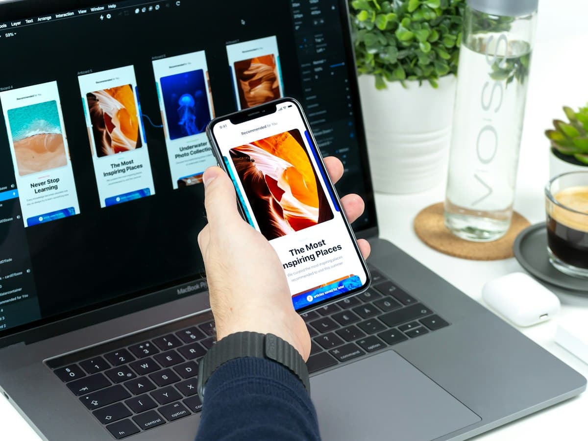 Designer reviewing a mobile app layout on a phone in front of a laptop showing screen designs